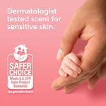 Hand holding baby's tiny hand with 'Safer Choice' seal and text about dermatologist-tested scent on pink background