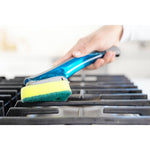 Hand using a Scotch-Brite dishwand to scrub a gas stove grate, close-up on the sponge head and metal grate