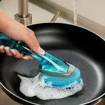 Hand holding a translucent blue Scotch-Brite dishwand scrubbing a nonstick frying pan with soap suds