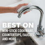 Hand holding a white dual-action Scotch-Brite Greener Clean pad cleaning a chrome faucet above a kitchen sink