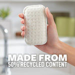 Hand holding a single beige-white Scotch-Brite Greener Clean pad in a bright kitchen with text about recycled content