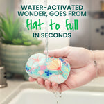 Hand holding a small floral pop-up sponge under running water with headline about the sponge expanding from flat to full.