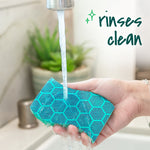 Hand holding a teal Scotch-Brite sponge under running faucet with overlay text "rinses clean" and kitchen background