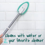 Scotch‑Brite scrubber head with green pad attached to a gray extendable handle scrubbing white subway tile wall.