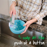 Person using a Scotch-Brite dishwand to wash a blue bowl over a kitchen sink with 'soap at the push of a button' text