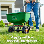 Person pushing a green Scotts spreader across a lawn, showing the spreader dispensing granules onto grass in front of a house.