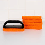 Orange scouring pads stacked beside a black handle scrubber on a kitchen counter with tiled backsplash.