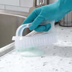 Hand wearing teal glove using white handled scrub brush to clean a countertop near a sink with suds