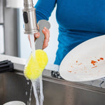 Lifestyle image of a person rinsing a yellow Dish Daddy sponge head under a kitchen faucet while holding a soiled plate over the sink.