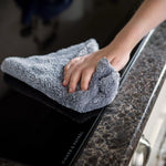 Hand wiping a black glass cooktop with a gray Scrub Daddy microfiber cloth on countertop