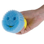 Hand holding a Scrub Daddy smiley-face blue scrub pad attached to a yellow hub, visible smile cutout