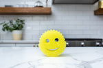 Yellow Scrub Daddy sponge placed on a marble countertop with a kitchen backsplash and stove softly out of focus behind it.