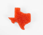 Loose orange Scrub Daddy sponge in the shape of Texas on a white background, showing textured open-cell foam