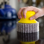 Hand pressing a yellow Scrub Hub with gray scrub pad on a glossy stovetop surface, reflected below