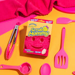 Pink Scrub Mommy sponge in its box surrounded by pink kitchen utensils and cloth on yellow surface