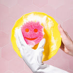 Gloved hand holding a yellow plate while another hand presents a pink Scrub Mommy sponge with soap suds against pink tile