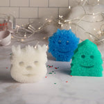 Lifestyle-style scene with three holiday-shaped scrubber sponges in white, blue and green on a countertop with string lights and baking props.