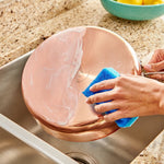 Hands using a blue scrubbing sponge to apply white cleaner to the exterior of a copper pan over a sink.