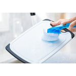 Hand scrubbing a white cutting board with a blue-handled brush under running water in a sink