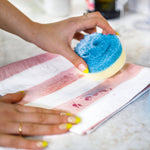 Hand scrubbing a red stain on a folded striped cloth with a round blue-and-yellow smiling-face sponge