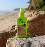 Shübug Active green spray bottle photographed on sunlit beach rocks with ocean and cliffs blurred in background.