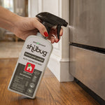 Hand holding and spraying a Shubug home pest control trigger bottle near a kitchen baseboard and oven.