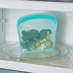 Teal reusable silicone bag filled with broccoli placed inside a microwave on the glass turntable