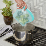 Hand holding a teal silicone food storage bag filled with broccoli over a pot of boiling water on a stovetop