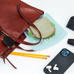 Teal reusable silicone sandwich bag with sandwich being placed beside a purse, phone, sunglasses, and pencils on a white surface