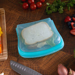 Teal reusable silicone sandwich bag with a sandwich inside placed on a wooden countertop with produce and a knife nearby
