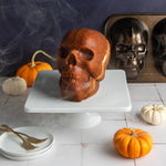Baked skull-shaped cake on a white pedestal with the Nordic Ware two-cavity skull pan visible in the background and small pumpkins.