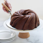 Person slicing a chocolate swirl bundt cake on a white cake stand, with clear lid visible in foreground