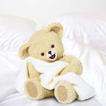 Snuggle teddy bear posed on a white bed holding a towel, photographed as a lifestyle image on a white background.