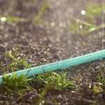 Close-up of a green soaker hose emitting a fine spray of water onto soil and grass, droplets visible on the hose