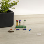 Product scene showing test tubes in a rack, colored reagent capsules, a pipette and soil next to a plant pot
