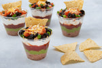 Four clear plastic cups used as snack or dip cups, styled with layered dip and tortilla chips on a light surface