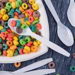White Solo plastic spoon resting over a bowl of colorful ring cereal with additional spoons arranged nearby on a dark wood surface.