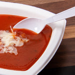 White Solo plastic spoon dipping into a bowl of tomato soup topped with shaved cheese on a wood table surface.