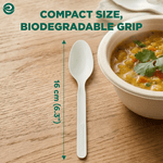 Single white compostable spoon laid on a wooden table with measurement arrows showing 16 cm and a bowl of soup at right
