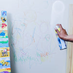 Hand spraying a foaming cleaner at a crayon-covered white wall next to stacked animal blocks.