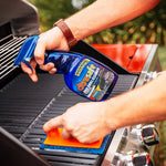 Person spraying Citrusafe BBQ Grill Cleaner onto grill while scrubbing with an orange-handled pad.