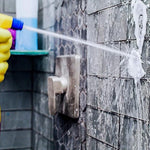 Hand in a yellow glove spraying foaming cleaner onto tiled shower wall, foam visible on grout and tile