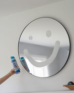 Hand holding a Sprayway glass cleaner can in front of a round mirror with a smiley face wiped into the condensation