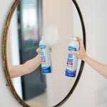 Hand spraying Sprayway glass cleaner can toward a round mirror, mirror reflecting the can and foam on the glass