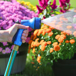Hand-held blue Sprout garden hose nozzle spraying a fine stream toward orange and pink flowers in a garden