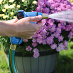 Hand holding a Sprout blue nozzle spraying water over pink flowers in a garden planter.