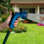Hand gripping a blue Sprout metal rear-trigger hose nozzle spraying strong water jets with a blurred house background