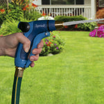 Hand-held blue Sprout hose nozzle spraying a narrow jet of water with a house and lawn blurred in background.