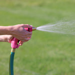 Two hands gripping a pink Sprout nozzle attached to a hose, producing a steady spray over grass
