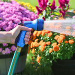 Person holding a blue Sprout rear-trigger nozzle spraying water over potted orange and pink blooms outdoors
